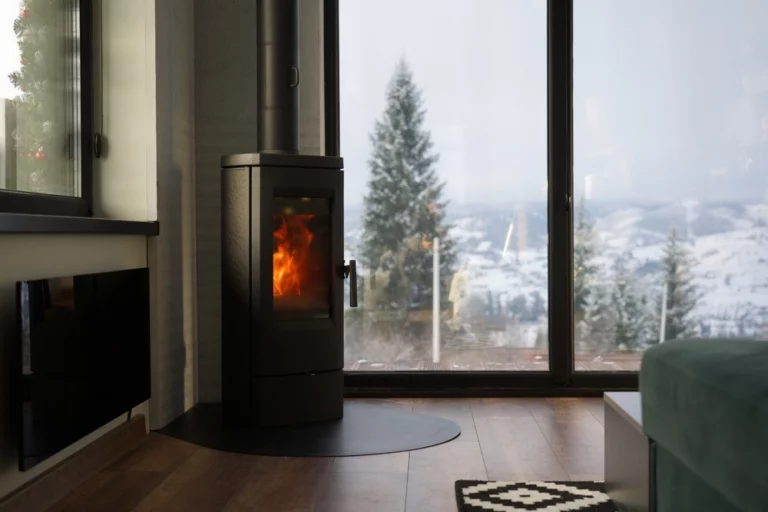 Moderner Kaminofen brennt im Wohnraum, große Fensterfront mit Winterlandschaft im Hintergrund.