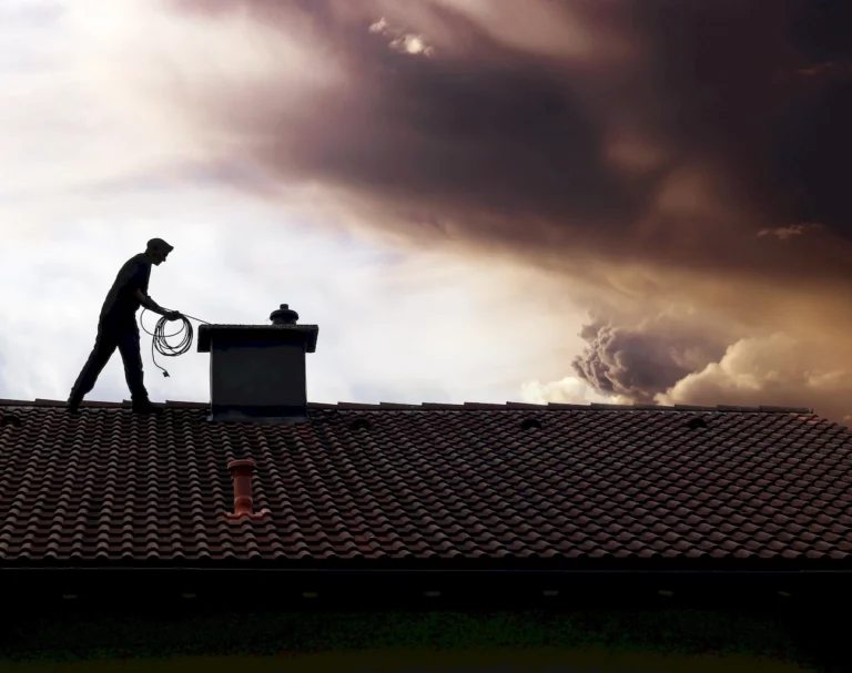 Schornsteinfeger auf einem Hausdach bei der Arbeit am Schornstein, mit Werkzeug in der Hand und dramatischem Himmel im Hintergrund.